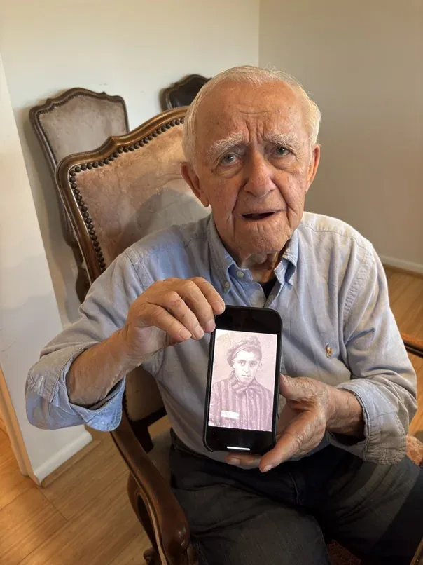 Arthur Gelbart holding a phone displaying a historical photograph of himself in prisoner clothing.