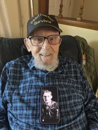 Ira Schab holding a phone displaying a photograph of himself in Navy uniform.